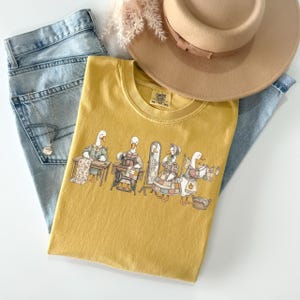May include: A mustard yellow t-shirt featuring a graphic of four geese engaged in sewing and laundry tasks, including quilting, using a sewing machine, looking in a mirror, and hanging clothes on a line. The shirt is paired with light blue denim jeans.