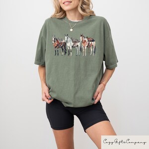 May include: A green t-shirt with a graphic of four horses with saddles. The horses are brown, white, and black.
