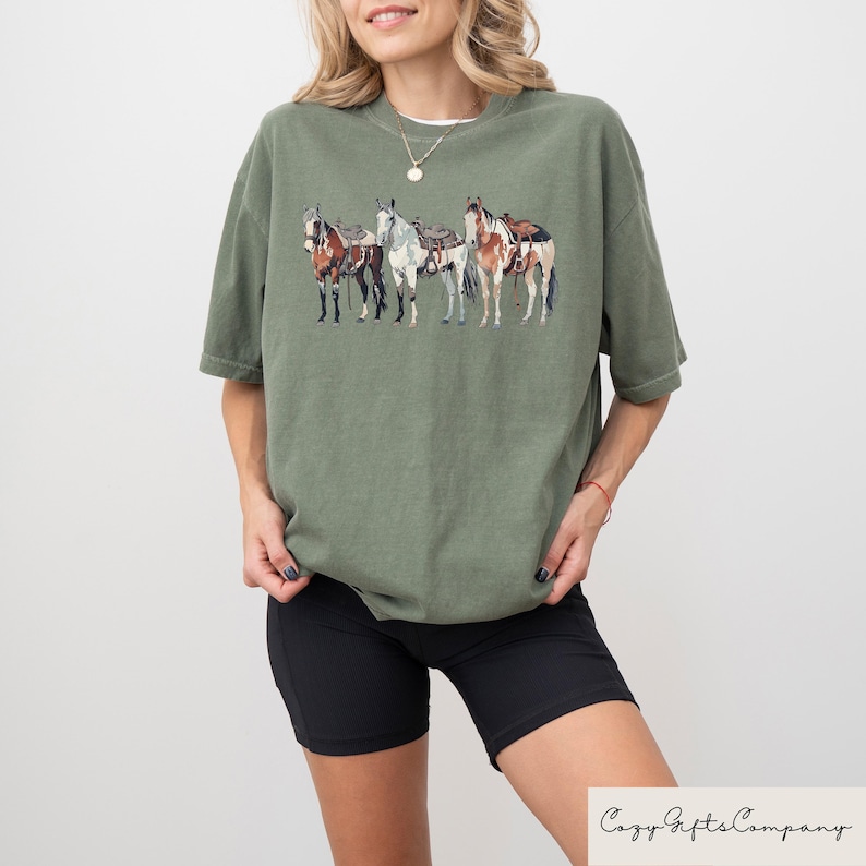 May include: A green t-shirt with a graphic of four horses with saddles. The horses are brown, white, and black.