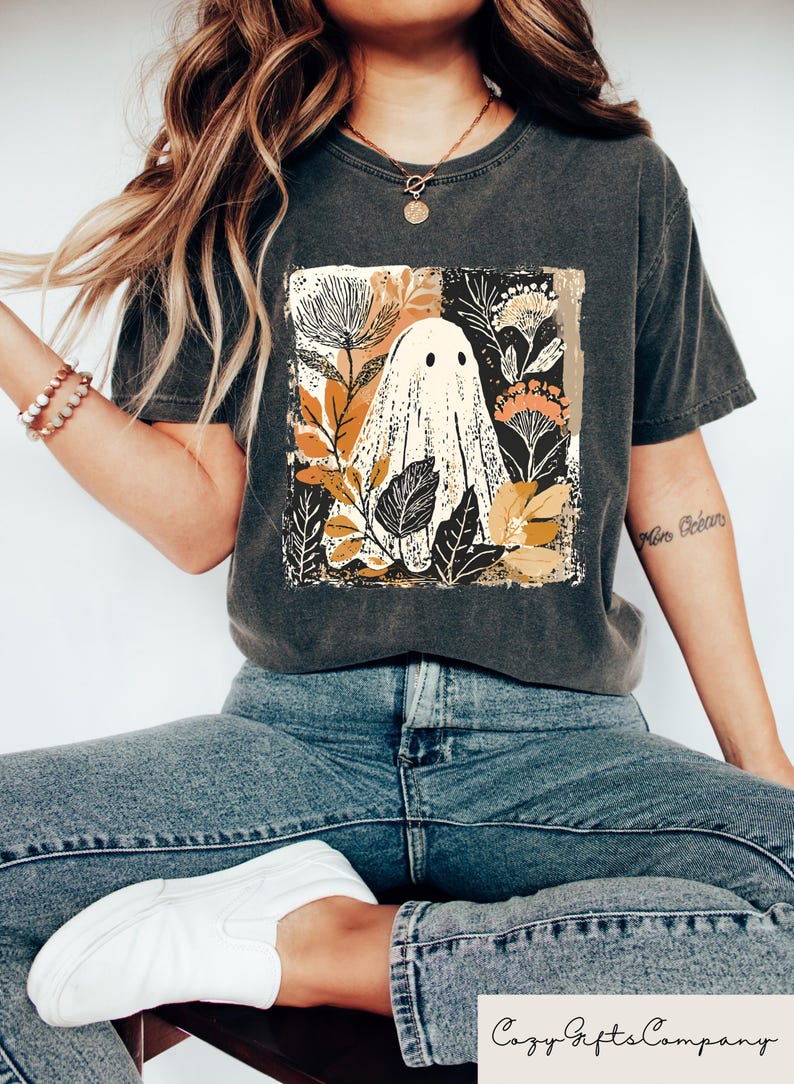 Op de afbeelding: Donkergrijs T-shirt met een vierkante afbeelding van een spook omringd door herfstbladeren en bloemen. Het spook is wit met zwarte ogen. Het T-shirt wordt gedragen met een blauwe jeans en witte schoenen. De tekst "Cozy Gifts Company" staat onderaan.