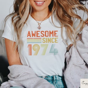 Pu&ograve; includere: Maglietta bianca con una grafica in stile vintage che dice "Awesome Since 1974" in arancione, giallo, verde e blu.