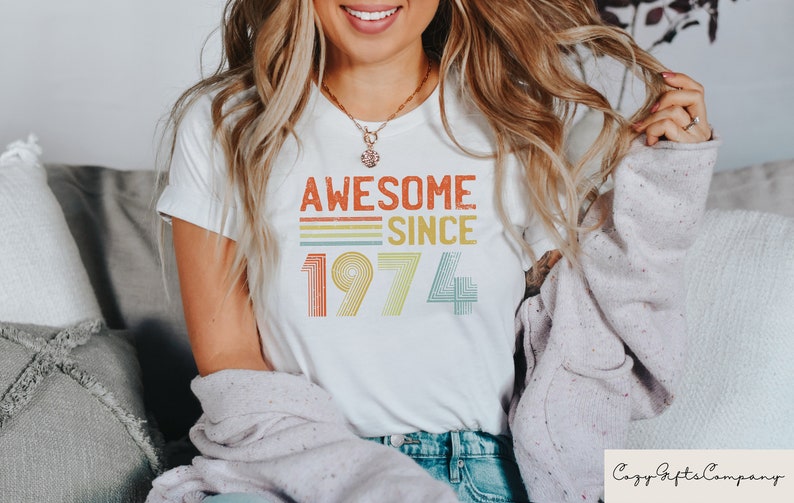 Pu&ograve; includere: Maglietta bianca con una grafica in stile vintage che dice "Awesome Since 1974" in arancione, giallo, verde e blu.