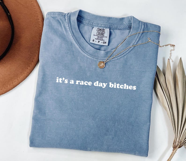 May include: A blue Comfort Colors t-shirt with the phrase "it's a race day bitches" in white lettering. A gold necklace with a pendant is draped across the shirt. A brown hat and dried palm leaves are in the background.