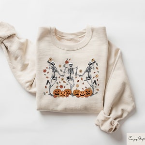 May include: A beige sweatshirt with a graphic print of three skeletons dancing with pumpkins and autumn leaves.