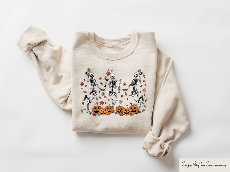 May include: A beige sweatshirt with a graphic print of three skeletons dancing with pumpkins and autumn leaves.