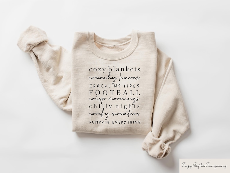 Op de afbeelding: Een beige sweatshirt met de tekst "cozy blankets, crunchy leaves, crackling fires, football, crisp mornings, chilly nights, comfy sweaters, pumpkin everything" in zwart gedrukt.