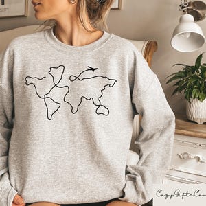 Puede incluir: Sudadera gris con un contorno negro del mundo y un peque&ntilde;o avi&oacute;n volando por encima.
