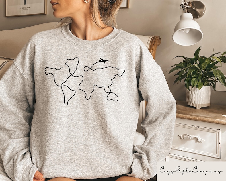 Puede incluir: Sudadera gris con un contorno negro del mundo y un peque&ntilde;o avi&oacute;n volando por encima.