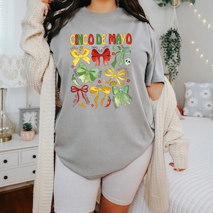 May include: A gray t-shirt with the text "Cinco de Mayo" printed on the front. The shirt features a design of colorful bows with various Mexican-themed words and images, including "Fiesta", "Chips & Salsa", "Tacos", "Spicy", "Hot Sauce", "Margaritas", "Queso", "Guacamole", "Mexican Street Corn", "Jalape&ntilde;o", "Lime", "Tomatillo", and "Serrano Pepper".