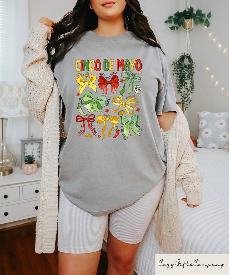 May include: A gray t-shirt with the text "Cinco de Mayo" printed on the front. The shirt features a design of colorful bows with various Mexican-themed words and images, including "Fiesta", "Chips & Salsa", "Tacos", "Spicy", "Hot Sauce", "Margaritas", "Queso", "Guacamole", "Mexican Street Corn", "Jalape&ntilde;o", "Lime", "Tomatillo", and "Serrano Pepper".
