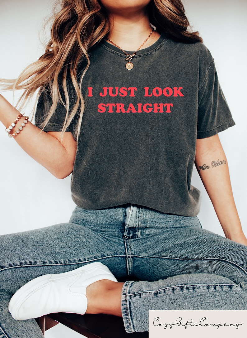 May include: A dark gray t-shirt with the text "I JUST LOOK STRAIGHT" in red lettering. The model is wearing the shirt with blue jeans and white sneakers.