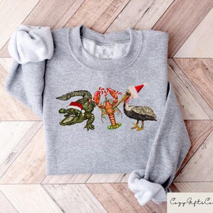 May include: A gray sweatshirt with a Christmas-themed design featuring a pelican wearing a Santa hat, a lobster, and an alligator wearing a Santa hat.