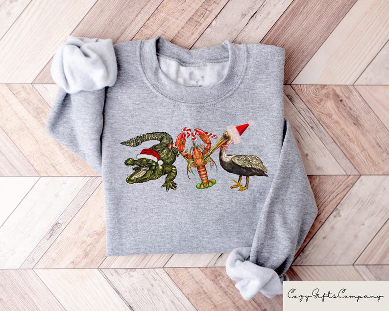 May include: A gray sweatshirt with a Christmas-themed design featuring a pelican wearing a Santa hat, a lobster, and an alligator wearing a Santa hat.
