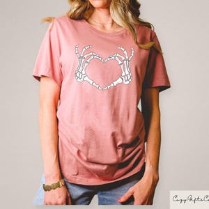 Może przedstawiać: R&oacute;żowy t-shirt z białym nadrukiem szkieletu dłoni tworzącym kształt serca. Nadruk głosi "Love You To Death".