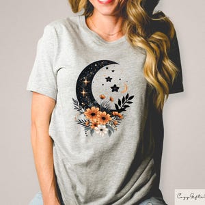 May include: A light gray t-shirt featuring a crescent moon design filled with stars and accented with orange and white flowers. The shirt has a crew neck and short sleeves. The text "Cozy Gifts Company" is in the bottom right corner.