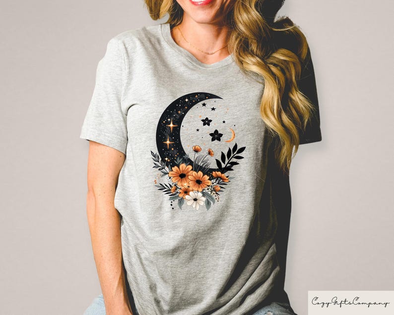 May include: A light gray t-shirt featuring a crescent moon design filled with stars and accented with orange and white flowers. The shirt has a crew neck and short sleeves. The text "Cozy Gifts Company" is in the bottom right corner.