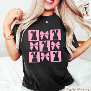 May include: Black t-shirt with a grid pattern of pink squares, each containing a black bunny silhouette or a pink bow. The shirt has a crew neck and short sleeves. The Cozy Gifts Company logo is visible.