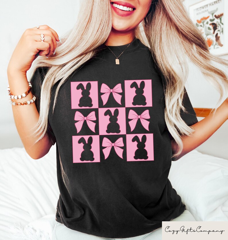 May include: Black t-shirt with a grid pattern of pink squares, each containing a black bunny silhouette or a pink bow. The shirt has a crew neck and short sleeves. The Cozy Gifts Company logo is visible.