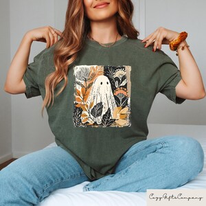 Op de afbeelding: Donkergroen T-shirt met een vierkante afbeelding van een spook omringd door herfstkleurige bloemen en bladeren. Het spook is wit met zwarte ogen. Het shirt heeft een relaxte pasvorm.
