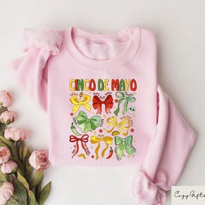 May include: Pink sweatshirt with a graphic design of colorful bows and the text "Cinco de Mayo".