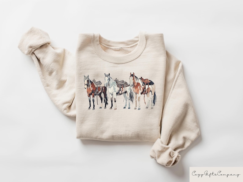 May include: A beige sweatshirt with a graphic print of four horses with saddles. The horses are brown, white, and black.