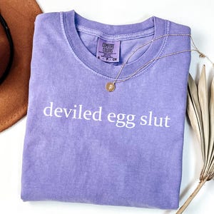 Puede incluir: Una camiseta lavanda Comfort Colors con la frase "deviled egg slut" impresa en blanco. Un collar dorado con un colgante descansa sobre la camiseta. La camiseta est&aacute; doblada y un sombrero marr&oacute;n es visible en el fondo.