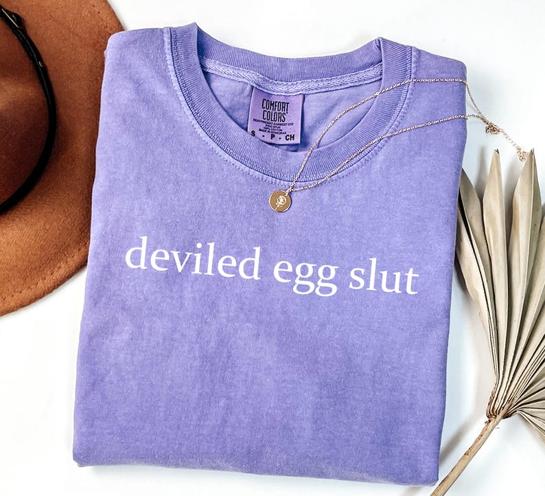 Puede incluir: Una camiseta lavanda Comfort Colors con la frase "deviled egg slut" impresa en blanco. Un collar dorado con un colgante descansa sobre la camiseta. La camiseta est&aacute; doblada y un sombrero marr&oacute;n es visible en el fondo.