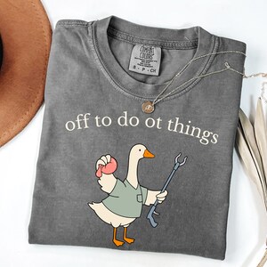 May include: A charcoal gray t-shirt featuring the white text "off to do ot things" above a cartoon goose wearing a sage green shirt. The goose holds a pink object in one wing and a blue reaching tool in the other. The shirt has a Comfort Colors label.