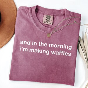 May include: A mauve Comfort Colors t-shirt with the text "and in the morning I'm making waffles" in white. A gold necklace with a pendant rests on the shirt. A brown hat and dried plant are in the background.