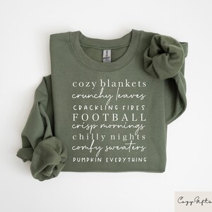 Op de afbeelding: Een groen sweatshirt met witte tekst die "cozy blankets, crunchy leaves, crackling fires, football, crisp mornings, chilly nights, comfy sweaters, pumpkin everything" luidt.