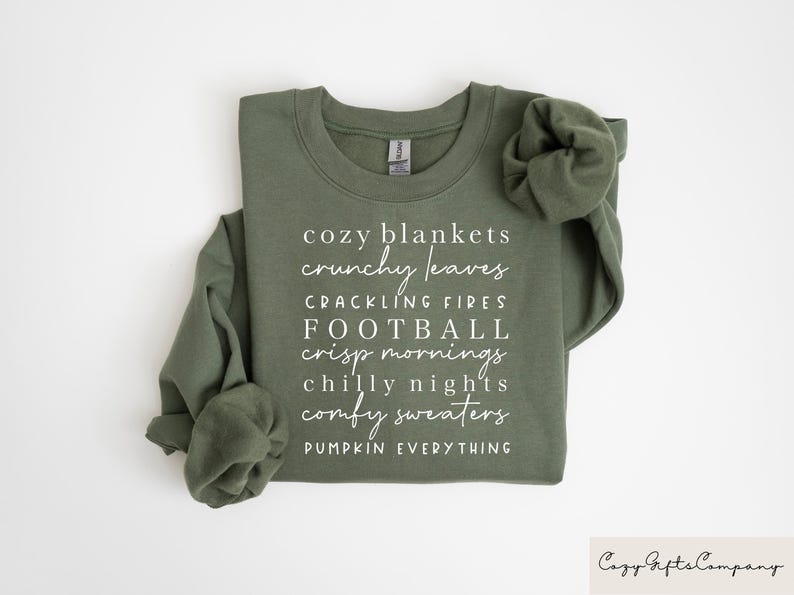 Op de afbeelding: Een groen sweatshirt met witte tekst die "cozy blankets, crunchy leaves, crackling fires, football, crisp mornings, chilly nights, comfy sweaters, pumpkin everything" luidt.