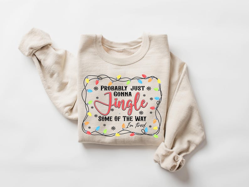 May include: A cream-coloured sweatshirt featuring the phrase "Probably Just Gonna Jingle Some of the Way I'm Tired". The design includes a border of colourful fairy lights and snowflakes, ideal for Christmas.