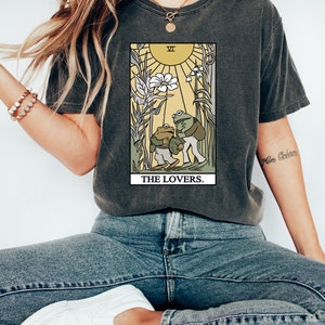 May include: A dark grey t-shirt featuring a graphic of the Lovers tarot card with two frogs. The card is surrounded by a yellow border and the text "THE LOVERS." is printed below the card.