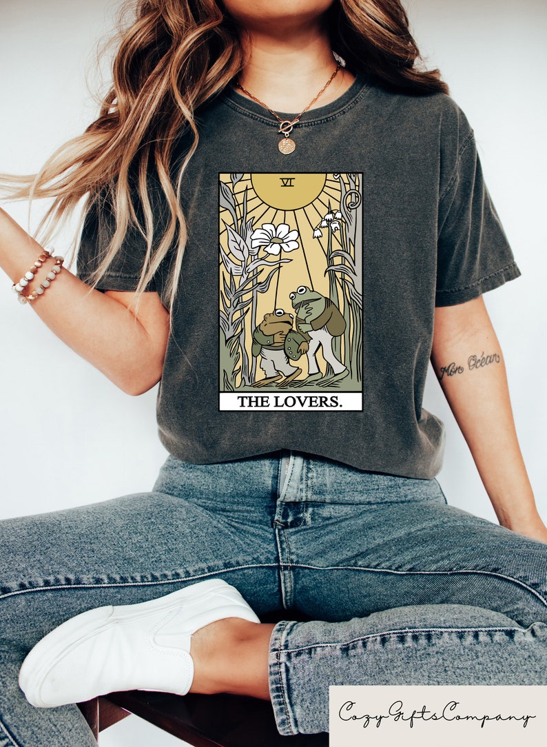 May include: A dark grey t-shirt featuring a graphic of the Lovers tarot card with two frogs. The card is surrounded by a yellow border and the text "THE LOVERS." is printed below the card.