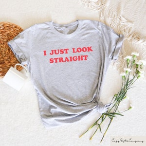 May include: Gray t-shirt with the text "I JUST LOOK STRAIGHT" in red.