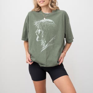 May include: Olive green t-shirt featuring a white graphic of a side profile of Jesus wearing a crown of thorns. The shirt has short sleeves and a relaxed fit. The model is wearing black shorts.