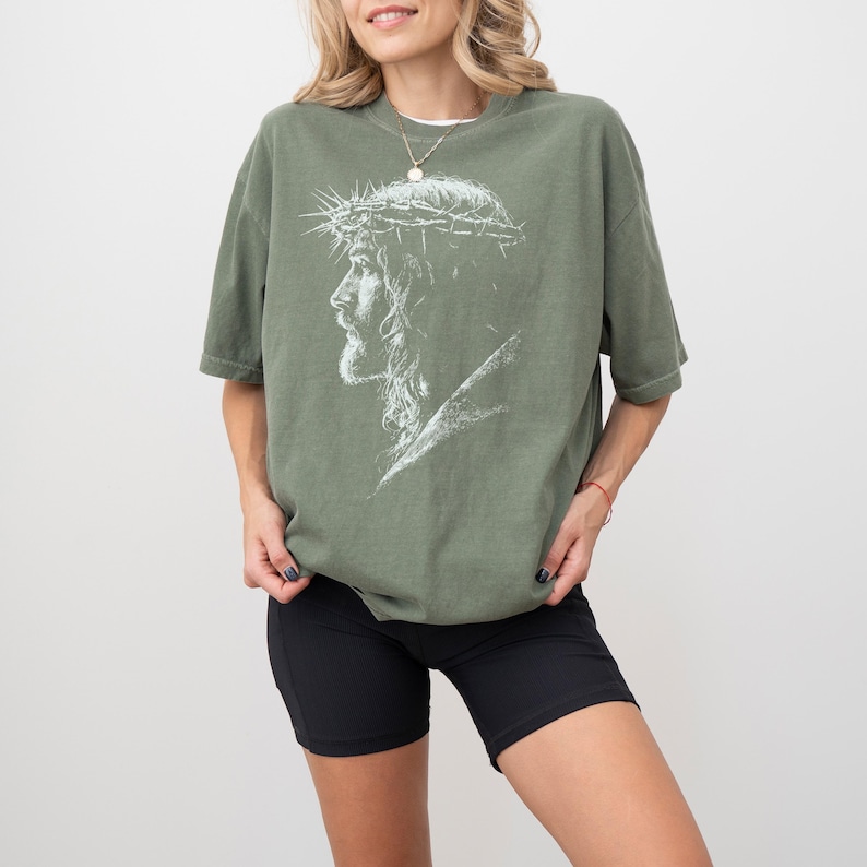 May include: Olive green t-shirt featuring a white graphic of a side profile of Jesus wearing a crown of thorns. The shirt has short sleeves and a relaxed fit. The model is wearing black shorts.