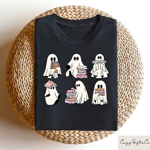Puede incluir: Camiseta negra con un gr&aacute;fico de seis fantasmas sosteniendo pilas de libros. Los fantasmas son blancos con ojos y bocas negros. Los libros son rosas, azules y blancos.
