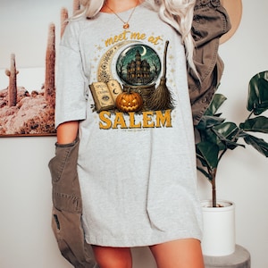 May include: A light gray t-shirt featuring a graphic print with the text "meet me at SALEM". The design includes a crystal ball containing a haunted house, an open book titled "SPELLS & CHARMS", a carved jack-o'-lantern, and a broomstick.