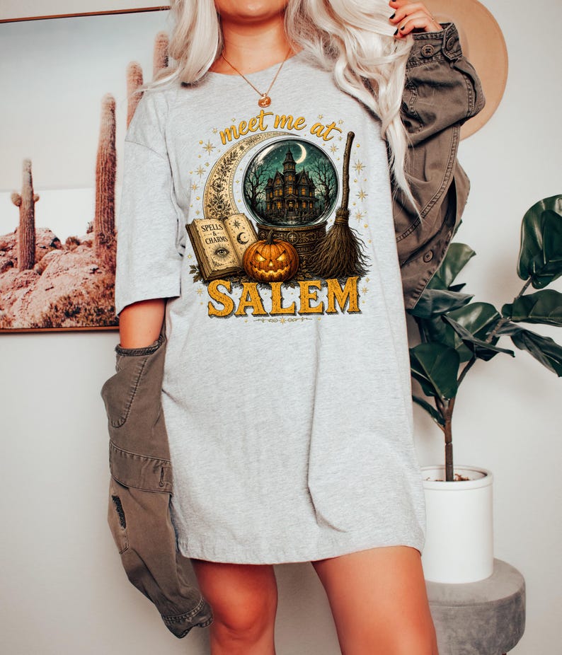 May include: A light gray t-shirt featuring a graphic print with the text "meet me at SALEM". The design includes a crystal ball containing a haunted house, an open book titled "SPELLS & CHARMS", a carved jack-o'-lantern, and a broomstick.