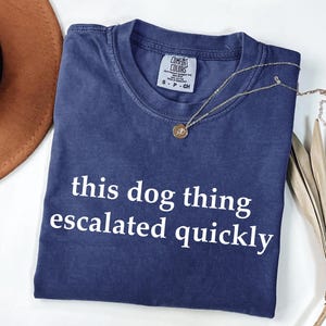 May include: A blue crew neck t-shirt with the white text "this dog thing escalated quickly" printed on the front. The shirt features a Comfort Colors brand label at the inner neckline, indicating a size small, and is made of 100% cotton.