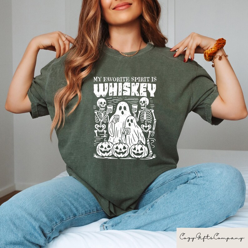 May include: Dark green t-shirt with the text "MY FAVORITE SPIRIT IS WHISKEY". The graphic features skeletons, ghosts, and jack-o'-lanterns. A fun, spooky design for Halloween. The shirt is made of a soft, comfortable material.