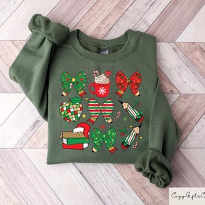 Puede incluir: Sudadera verde con un dise&ntilde;o navide&ntilde;o con lazos rojos y verdes, l&aacute;pices, una manzana, una taza de chocolate caliente y una pila de libros.