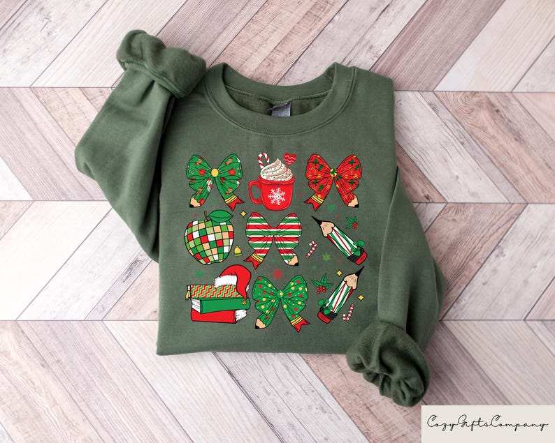 Puede incluir: Sudadera verde con un dise&ntilde;o navide&ntilde;o con lazos rojos y verdes, l&aacute;pices, una manzana, una taza de chocolate caliente y una pila de libros.