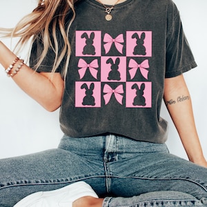May include: Dark grey t-shirt with a grid of pink squares, each containing a bunny or bow silhouette. The top has a crew neck and short sleeves. The text "Cozy Gifts Company" is visible at the bottom of the image.