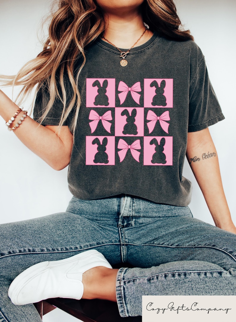 May include: Dark grey t-shirt with a grid of pink squares, each containing a bunny or bow silhouette. The top has a crew neck and short sleeves. The text "Cozy Gifts Company" is visible at the bottom of the image.