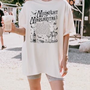 Puede incluir: Una camiseta blanca con un estampado gr&aacute;fico negro de una coctelera, una copa de martini y una botella de licor. El gr&aacute;fico tambi&eacute;n incluye el texto "Midnight Margaritas" y una lista de ingredientes para una poci&oacute;n m&aacute;gica.