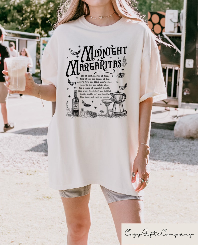 Puede incluir: Una camiseta blanca con un estampado gr&aacute;fico negro de una coctelera, una copa de martini y una botella de licor. El gr&aacute;fico tambi&eacute;n incluye el texto "Midnight Margaritas" y una lista de ingredientes para una poci&oacute;n m&aacute;gica.