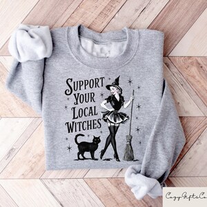 May include: A gray sweatshirt with the text "Support Your Local Witches" and a black and white illustration of a witch with a black cat and a broom.