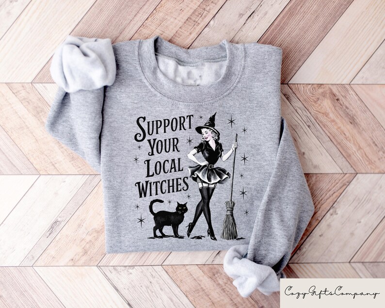 May include: A gray sweatshirt with the text "Support Your Local Witches" and a black and white illustration of a witch with a black cat and a broom.